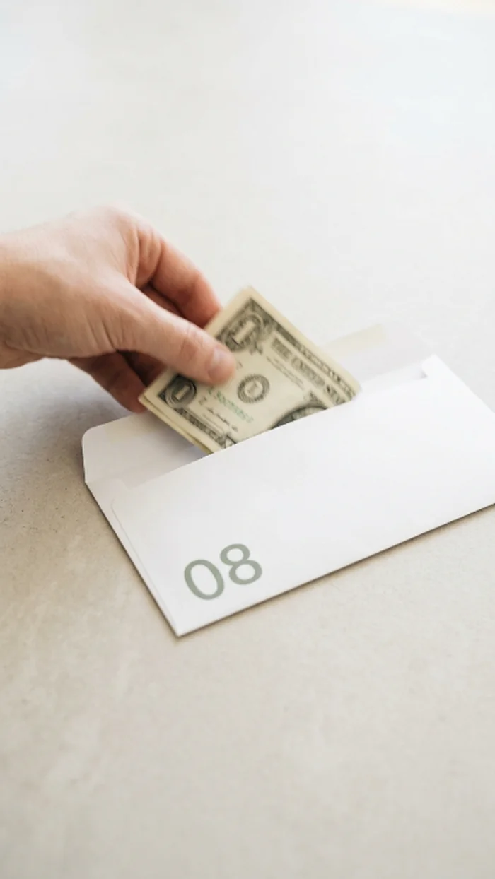 A hand tucking a folded dollar bill into a numbered paper envelope marked 08
